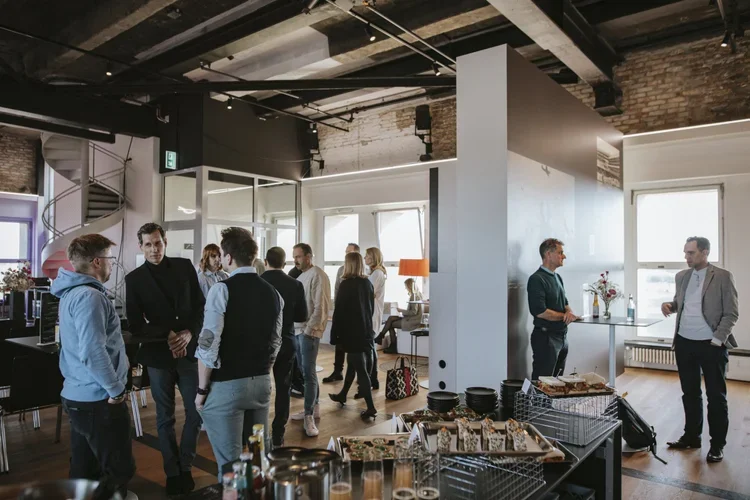 Group of people socializing and chatting in a modern, industrial-style office or event space with large windows and minimal decor.