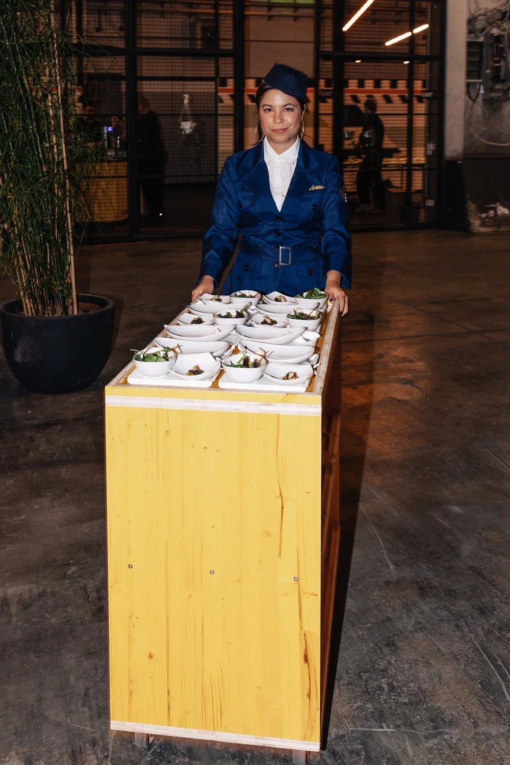 A woman dressed in a blue uniform and hat stands behind a yellow cart with multiple bowls of food, likely in a restaurant or catering setting.