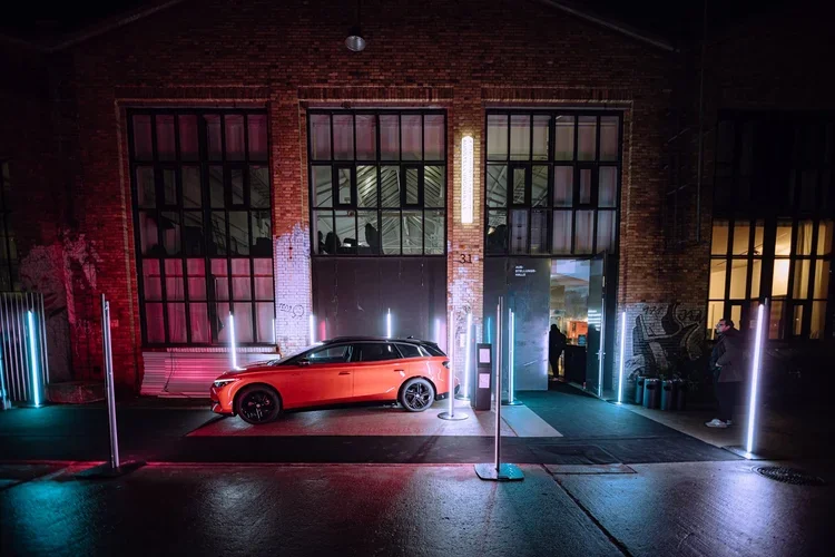 A red car parked outside a brick building with large windows at night, illuminated by colorful neon lights and vertical light fixtures.