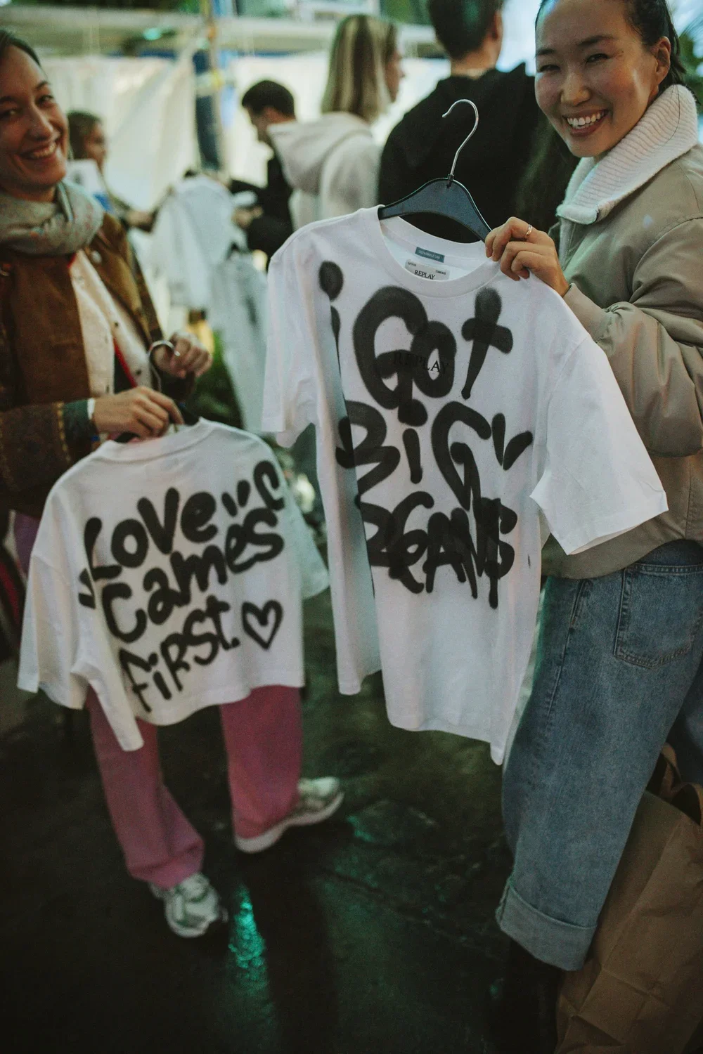 Two women shopping at a market, holding white T-shirts with bold black graffiti-style text. One shirt says 'Love's Camels First' with a heart, the other says 'Got Big Pals'.