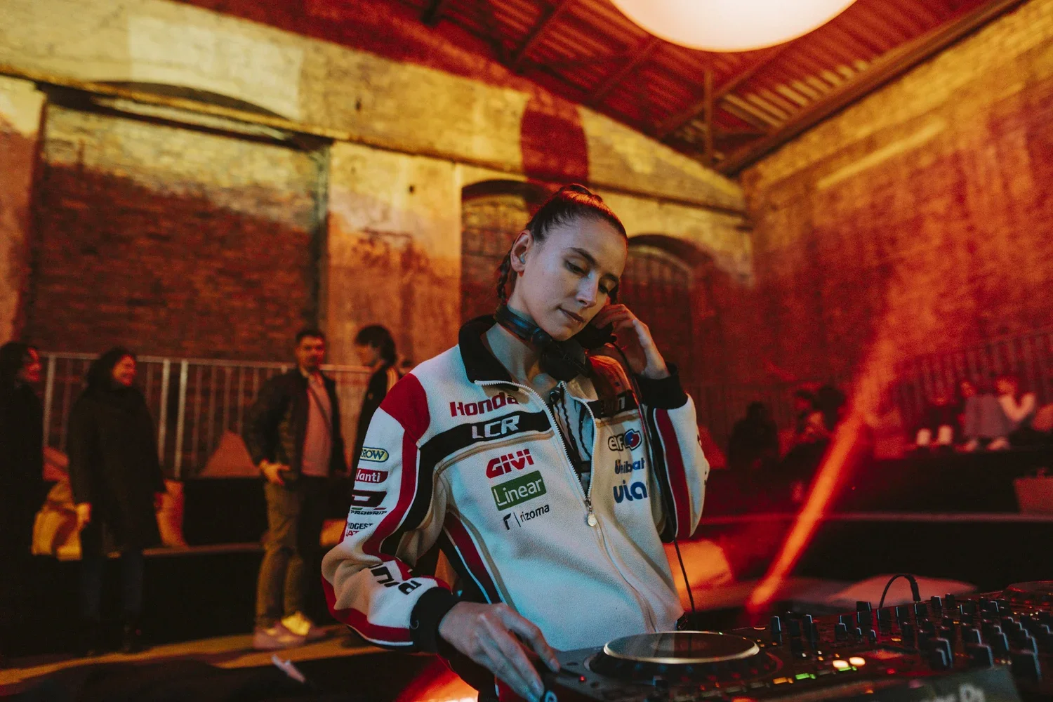 A female DJ or music mixer wearing a racing jacket with logos, using DJ equipment at a lively nightclub or event with colorful lighting and people in the background.