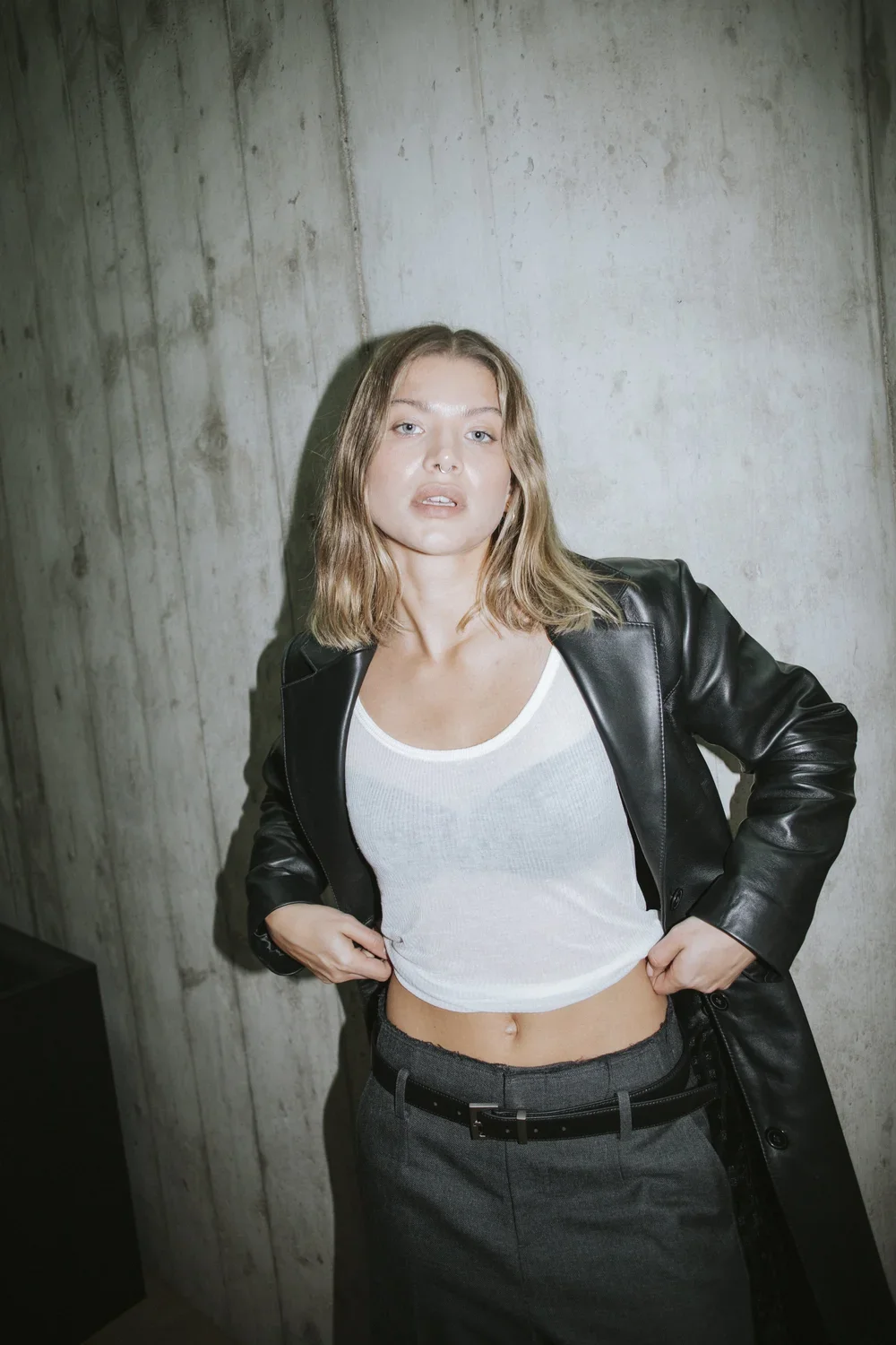 A woman with shoulder-length light brown hair, wearing a black leather jacket over a white cropped t-shirt and dark pants, standing against a concrete wall.