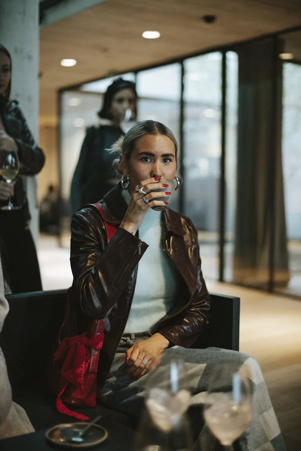 A young woman with blonde hair, hoop earrings, and a leather jacket is sitting at a table, holding a glass to her lips, surrounded by other people in a modern indoor setting.