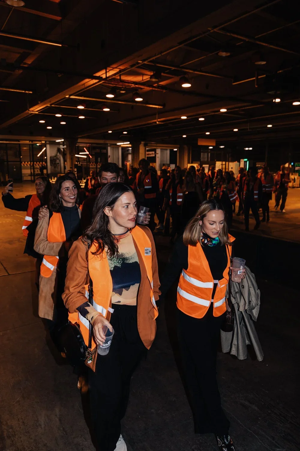 Group of people wearing orange safety vests walking through an indoor parking garage at night.