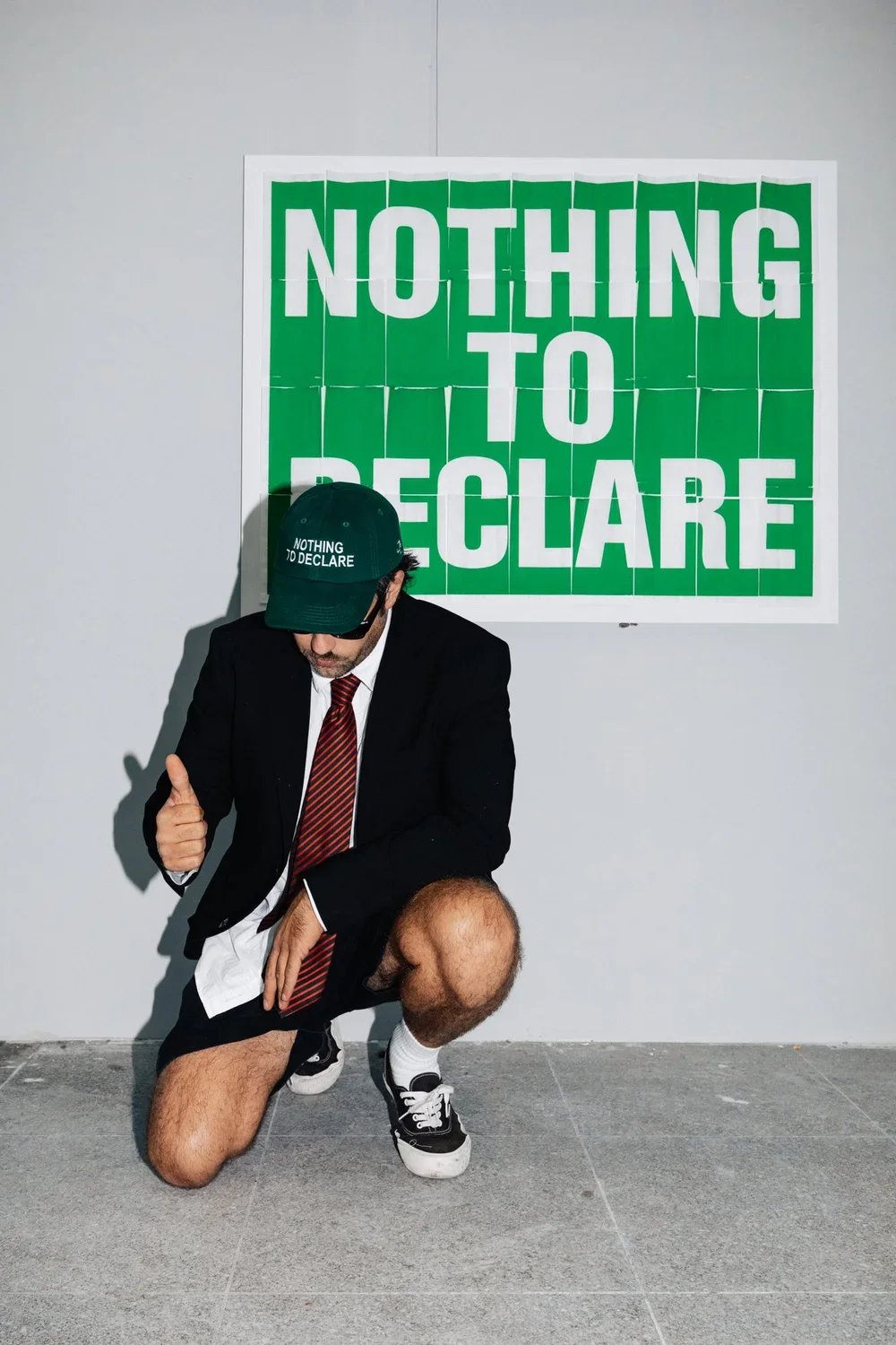 A man in a suit, shorts, and sneakers kneeling on one knee, giving a thumbs-up gesture. He is wearing a hat with the text 'Nothing to Declare'. Behind him, there is a green and white sign that says 'Nothing to Declare' in large letters.