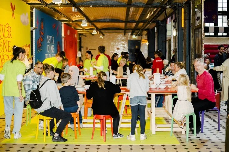 People gathered in a colorful, modern indoor space, sitting around tables, with some standing, engaged in dining or conversation, with vibrant walls and decor.