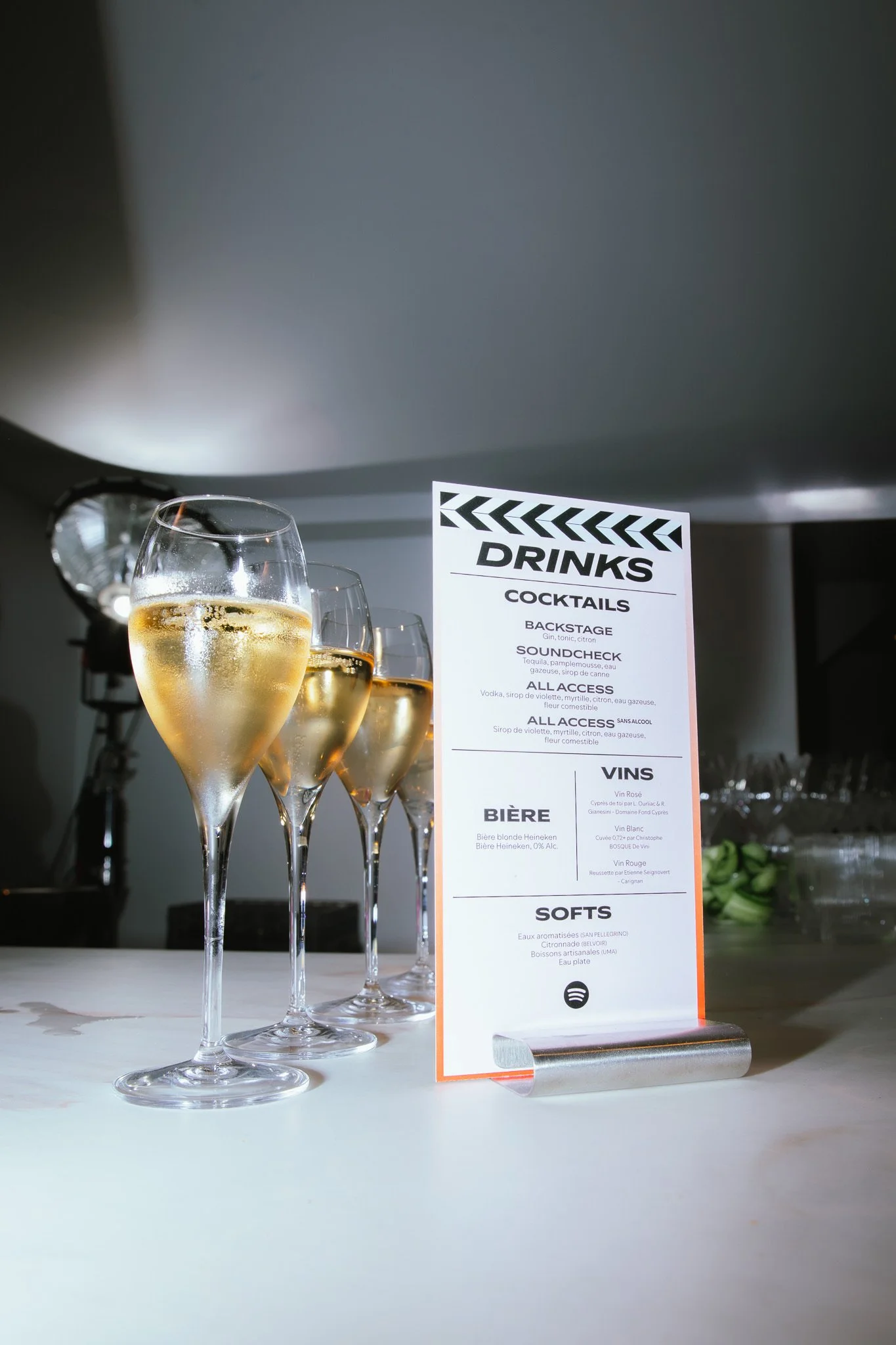 Four champagne glasses filled with a golden bubbly drink, placed in a row on a white surface with a drinks menu standing behind them.