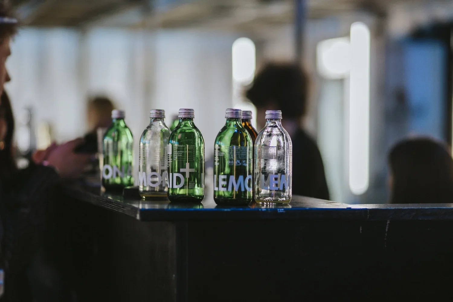Several glass bottles labeled with different flavors such as mint and lemon on a bar counter, with a blurred background of people and a dimly lit indoor setting.