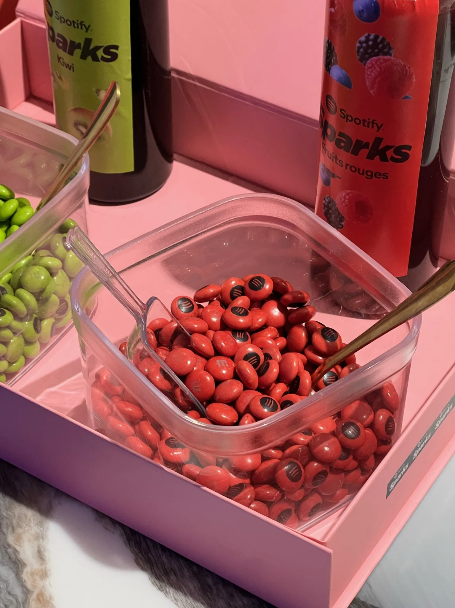 A plastic container of red jelly beans with black logos, alongside other containers of green jelly beans, set on a pink surface with beverage bottles in the background.