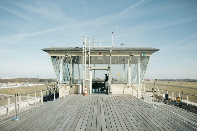 View of an airport control tower made of glass and metal, with a person inside, on a cloudy day. The tower stands on a wooden deck with safety railings, overlooking a flat landscape and runway.
