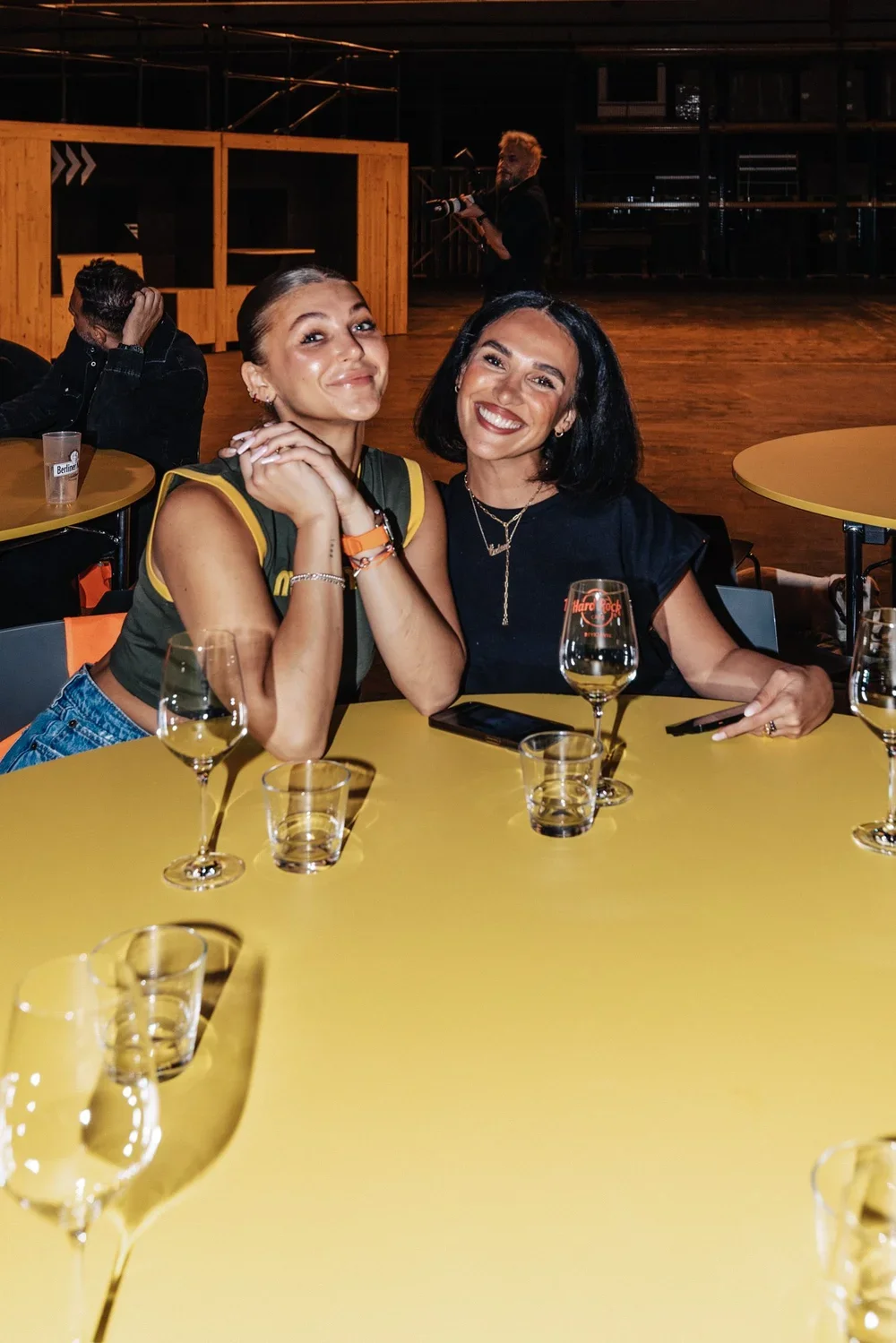 Two women smiling and holding hands at a yellow table with wine glasses, in a dimly lit lounge or bar setting.