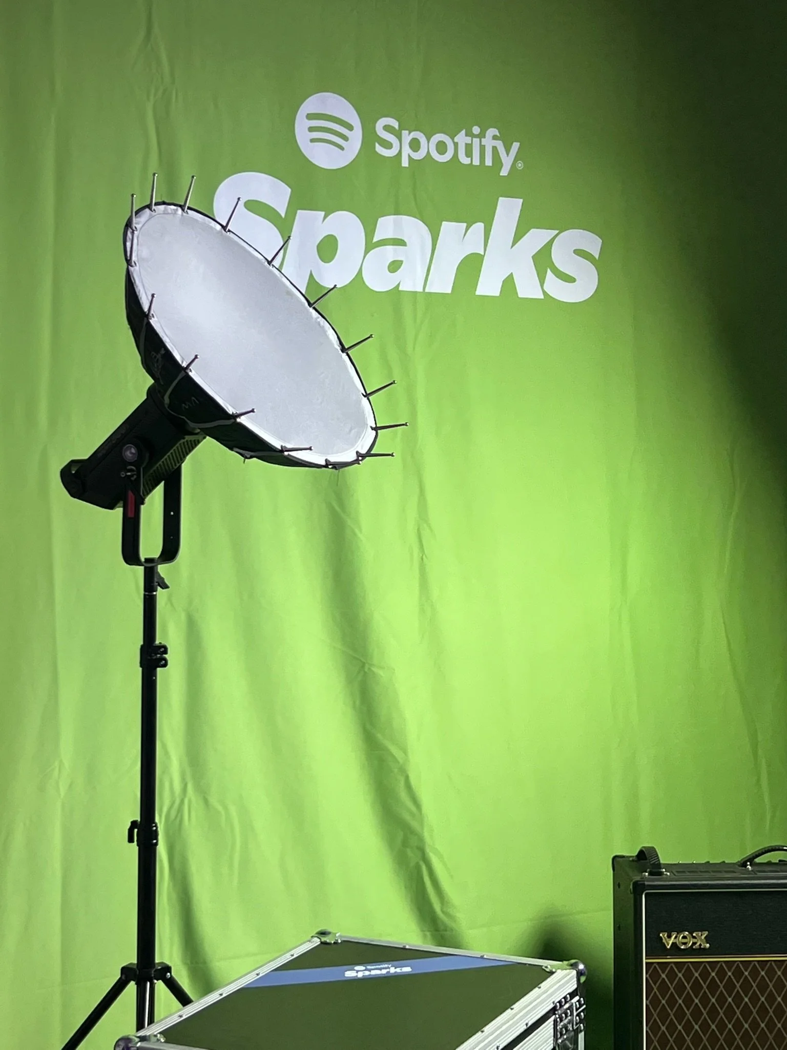 A photography setup with a green backdrop featuring the 'Spotify Spark' logo, a large professional studio light with a diffusing shade, a black case with a 'Spotify Spark' logo, and a small Vox amplifier.