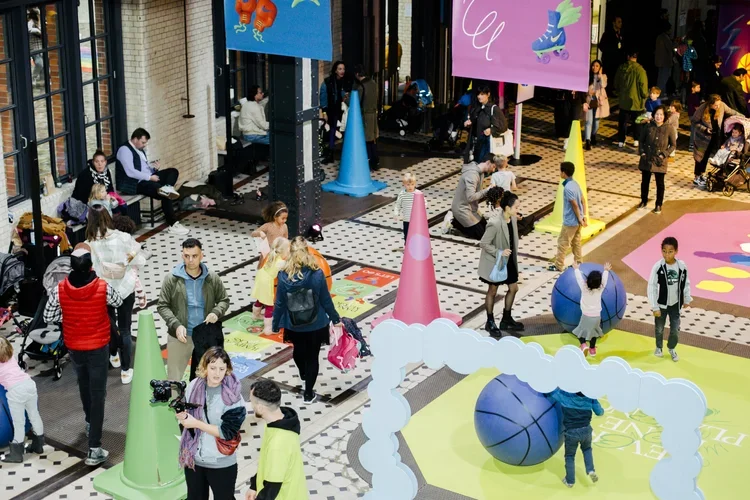 Children and adults gathered in an indoor play area with colorful cones, large balls, and playful decorations.