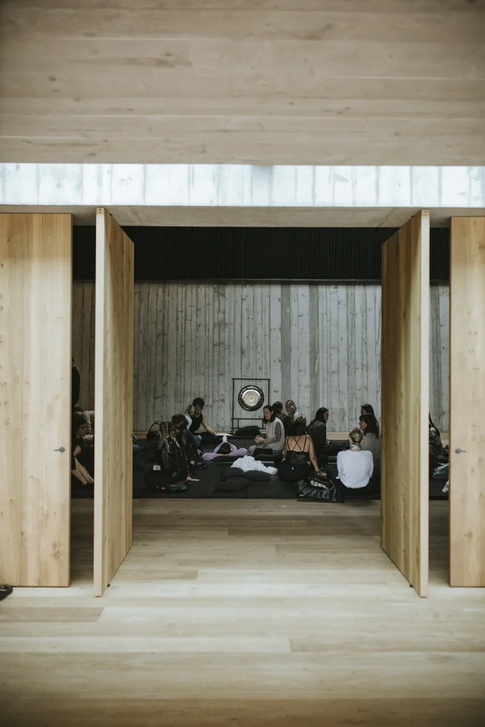 Group of people sitting on black cushions in a minimalist room with wooden walls and a large gong at the back.