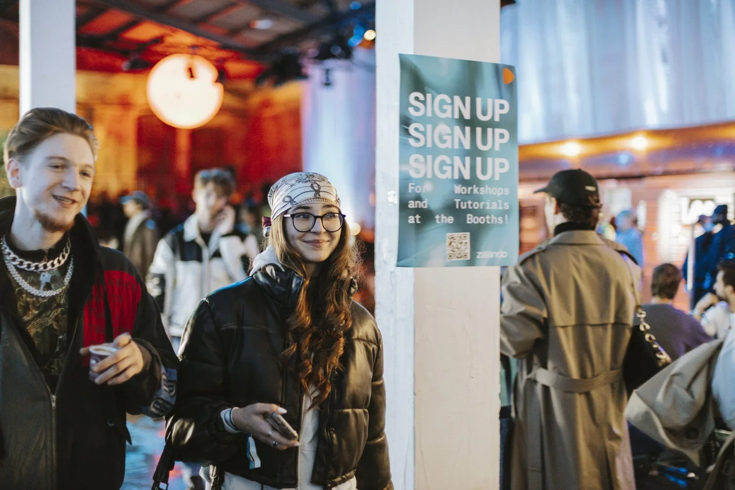People attending an event at a booth with a sign that reads 'Sign Up for Workshops and Tutorials at the Booths!'. The setting appears to be a busy indoor venue with a warm, colorful ambiance, and multiple individuals engaged in conversations.