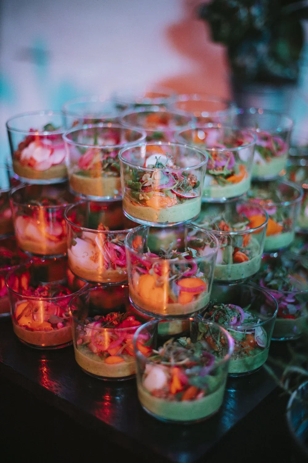Multiple small glass cups filled with colorful food, arranged on a dark surface, with blurred colorful lights in the background.