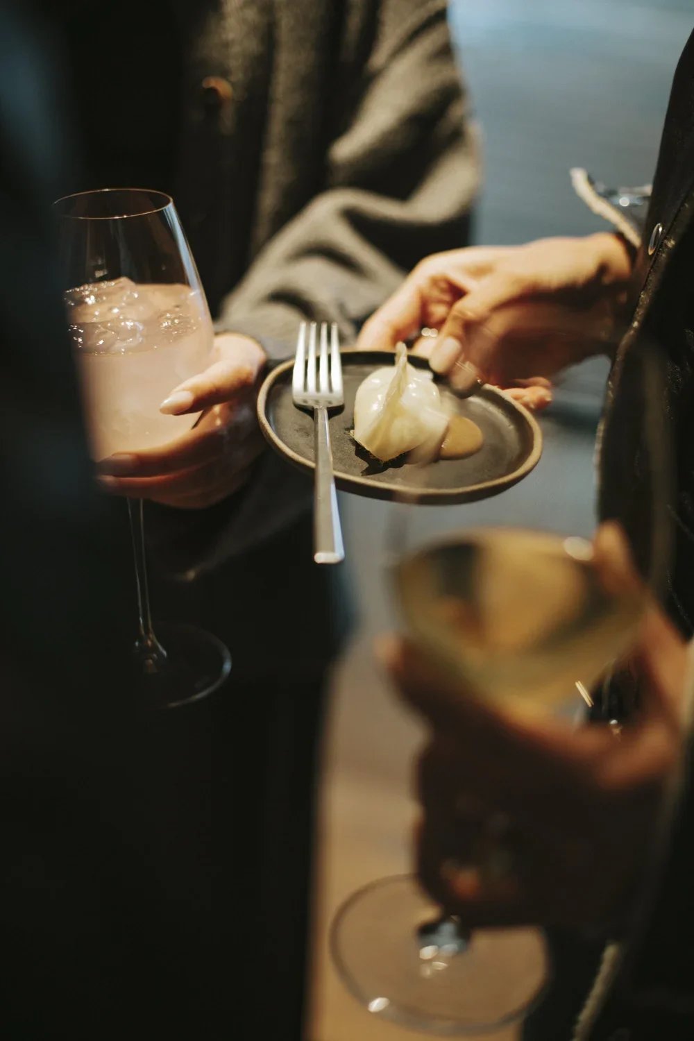 People holding glasses of drinks and a small plate with a dumpling, fork, and sauce in a social setting.