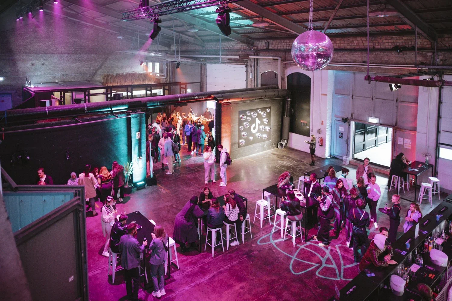 Interior of a lively event space with colorful lighting, a disco ball, a TikTok logo on the wall, people socializing and waiting in line for the bar, and a dance floor marked on the floor.