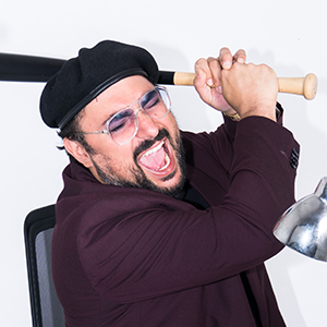 Man with glasses and a black hat holding a baseball bat over his shoulder, smiling widely.