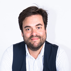 A man with dark hair and a beard smiling, wearing a light blue striped shirt and a dark vest, against a plain white background.