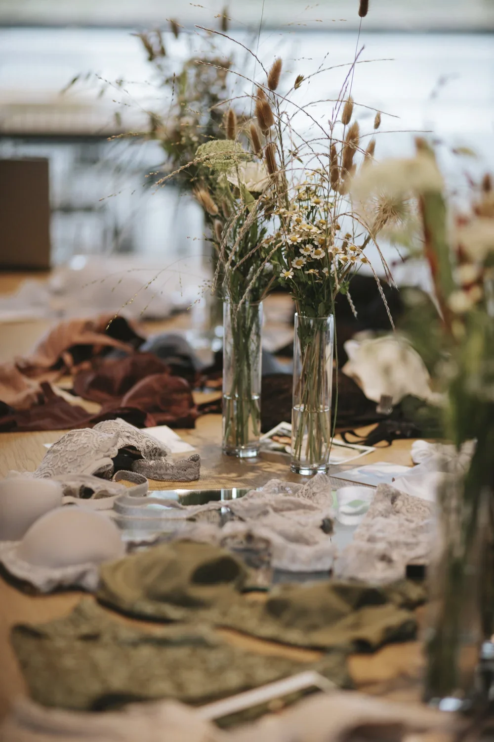 A table filled with various lace and fabric items, with three clear glass vases holding dried floral arrangements, including brown grasses and small white flowers.