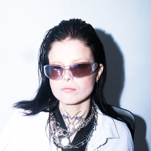 A woman with black hair wearing sunglasses, a light-colored shirt, and layered necklaces.