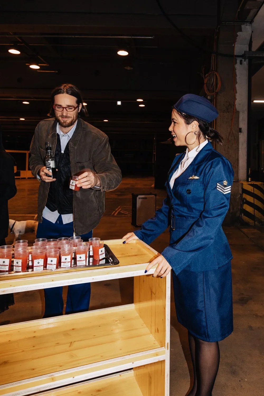 A woman in an airline uniform serving drinks at a table with small cups of tomato juice in an industrial-style interior.