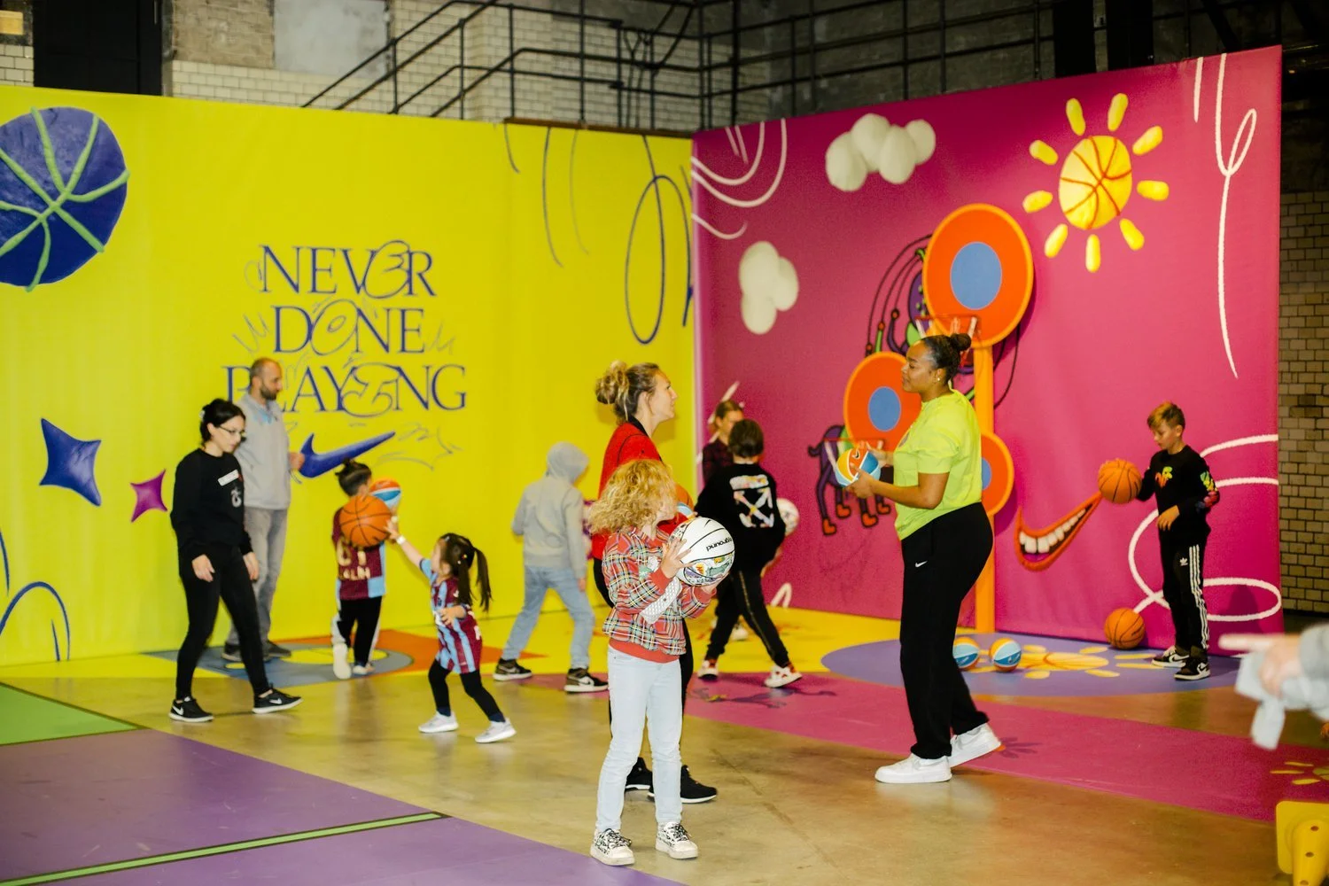 Children and adults at a colorful indoor basketball activity area, some children holding basketballs, with vibrant wall murals and motivational quote 'Never Done Playing' in the background.