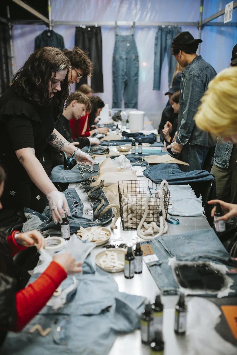 People working on denim clothing project at a long table in a tent or makeshift workspace, assembling or customizing jeans with various tools and supplies.