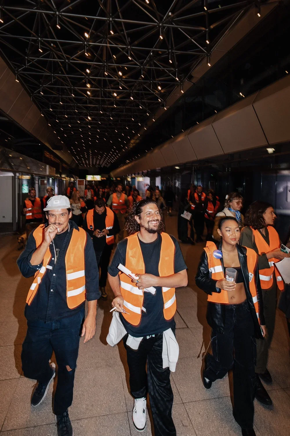 Group of people wearing orange safety vests walking through an airport corridor at night.