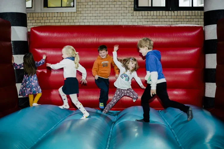 Five children playing on an inflatable bounce house with a red wall and striped columns.