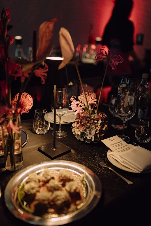 Elegant event table setting with floral centerpieces, wine glasses, and a menu, with a blurred background of people and dim lighting.