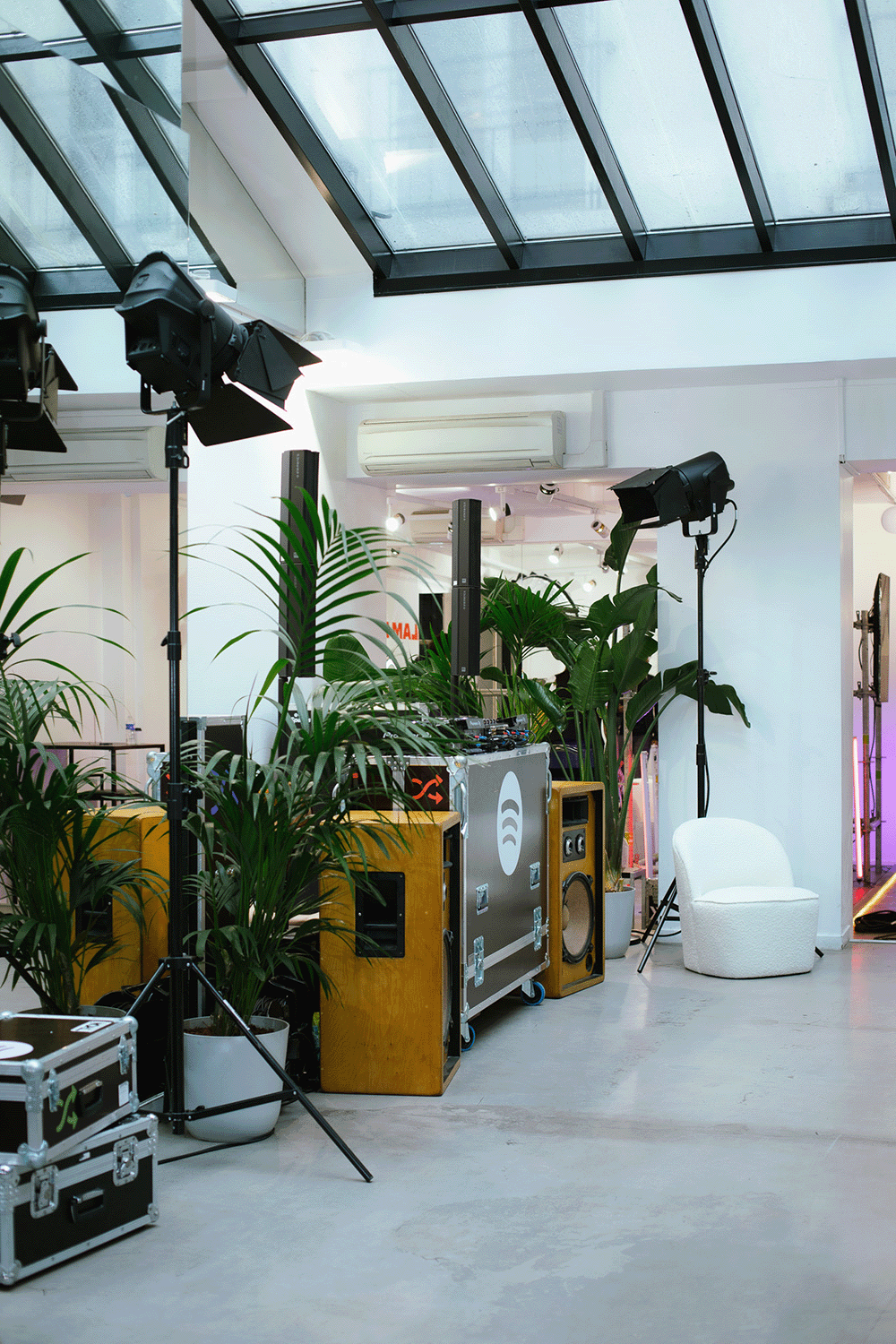Indoor event space with DJ equipment, large green plants, professional lighting, white seating, and a glass ceiling.
