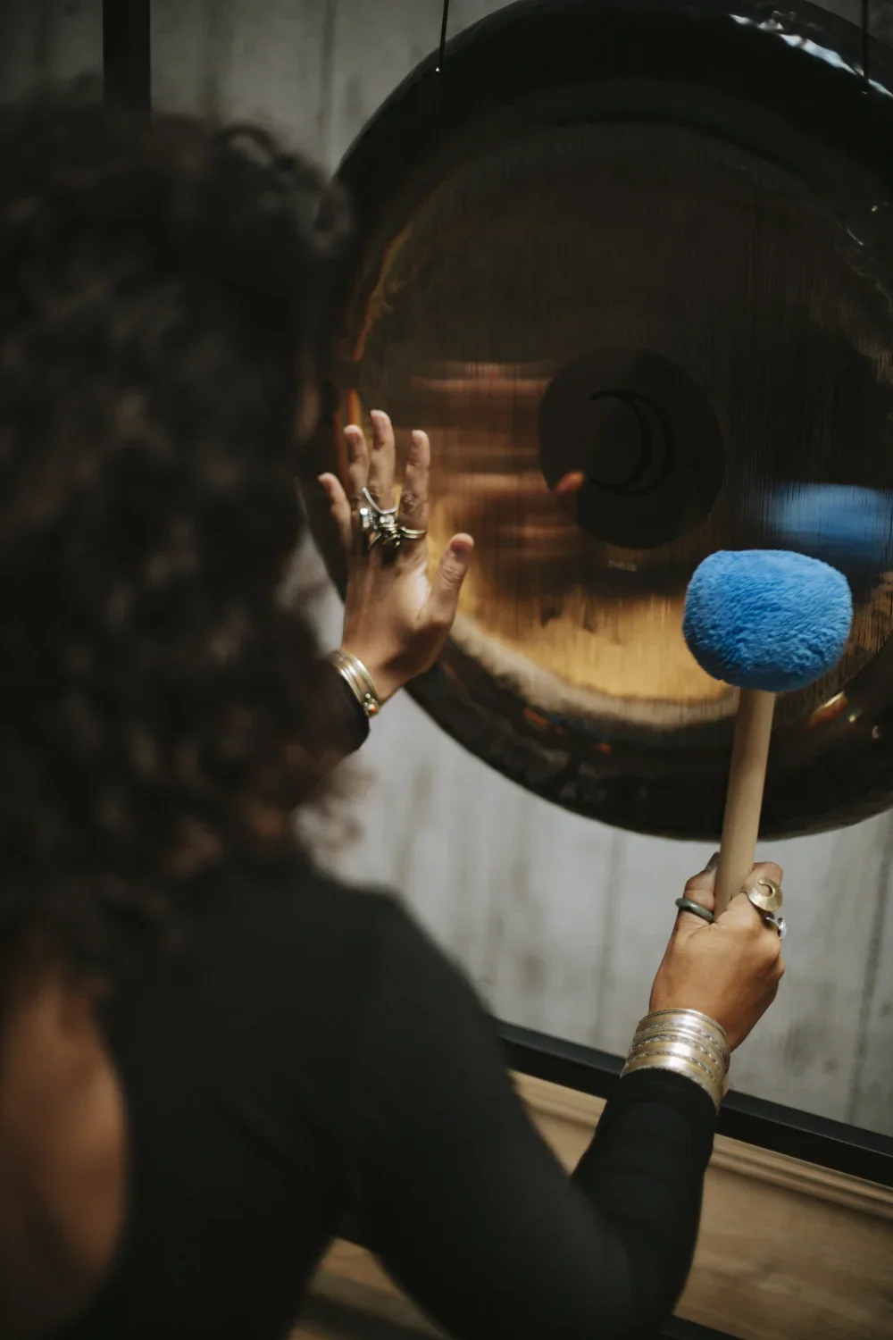A woman with curly hair and rings is hitting a large gong with a blue mallet.