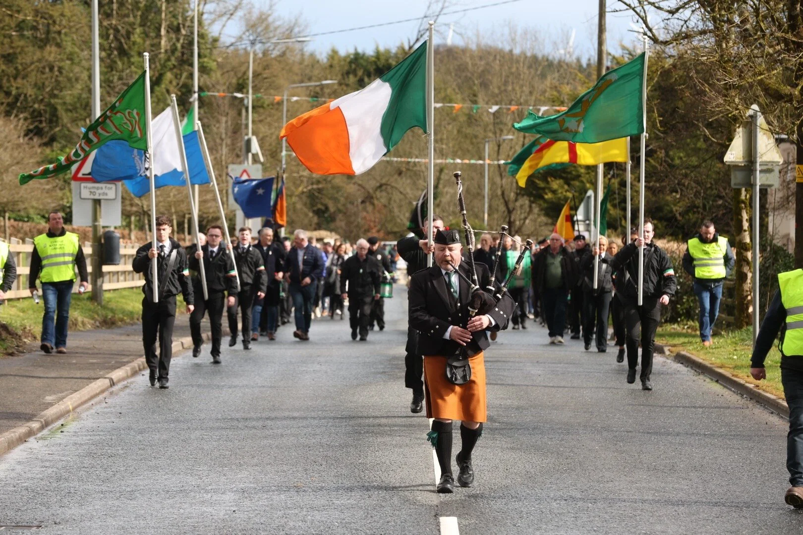 A parade featuring a man playing bagpipes, dressed in traditional Scottish attire, leading a group of people carrying various flags, including the Irish flag. The parade is walking on a street with trees in the background and some spectators and security personnel on the sides.