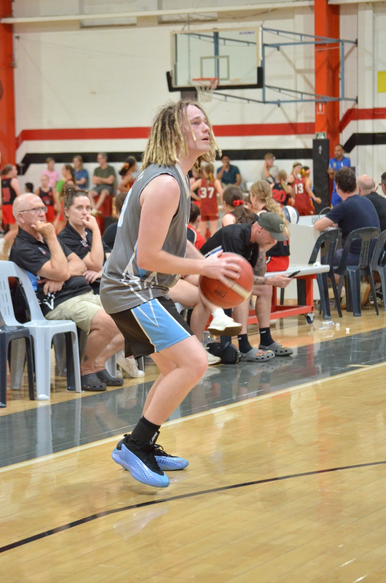 A young male basketball player with dreadlocks, wearing a gray uniform and black shoes with blue soles, is holding a basketball and preparing to make a shot during a game in a school gymnasium. Background shows seated spectators and a team sitting on the sideline, with other players and coaches in the scene.