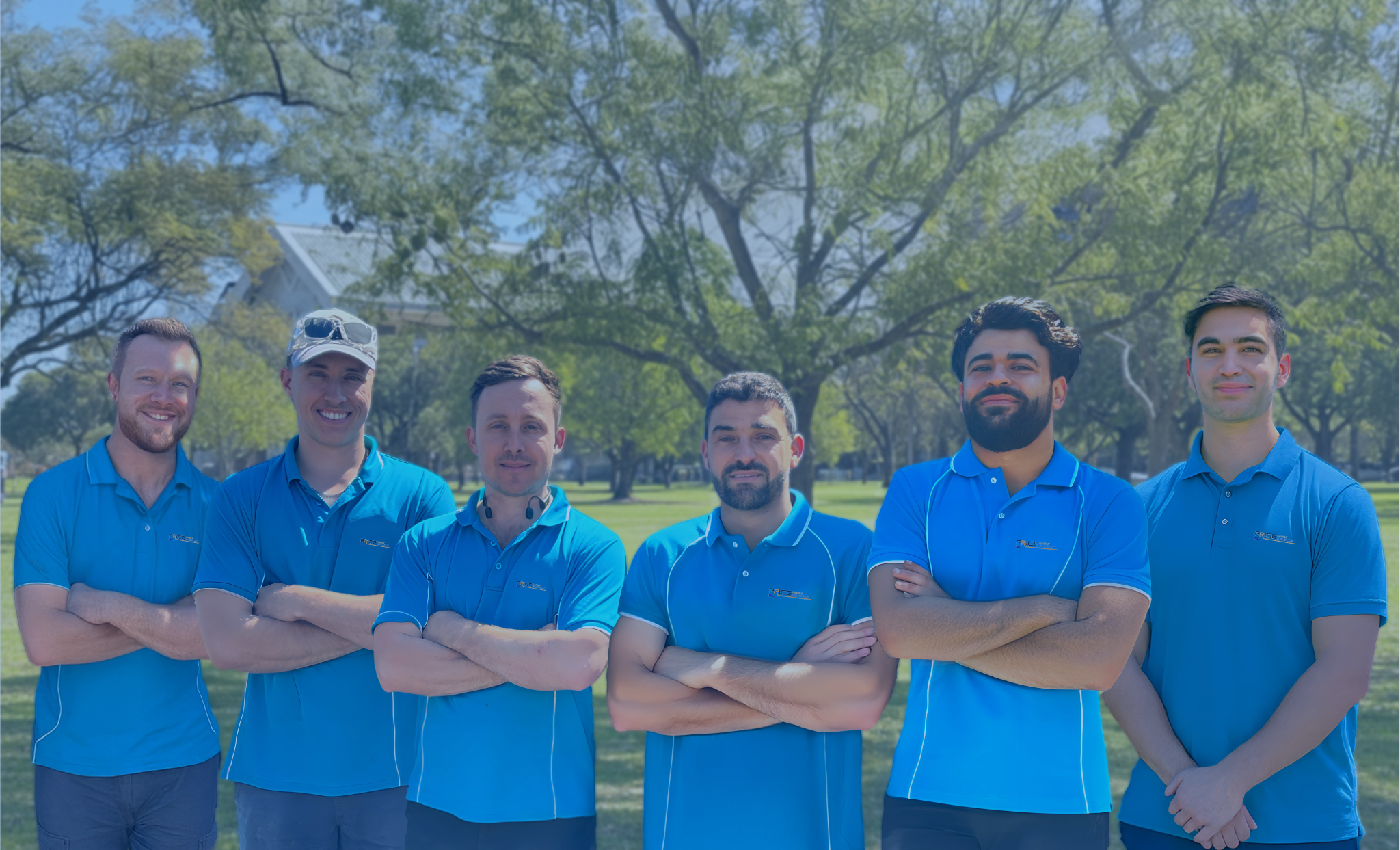 Group of six electricians from Price Family Electrical standing outside in a park, all wearing matching blue polo shirts with a logo, and standing in front of green trees.