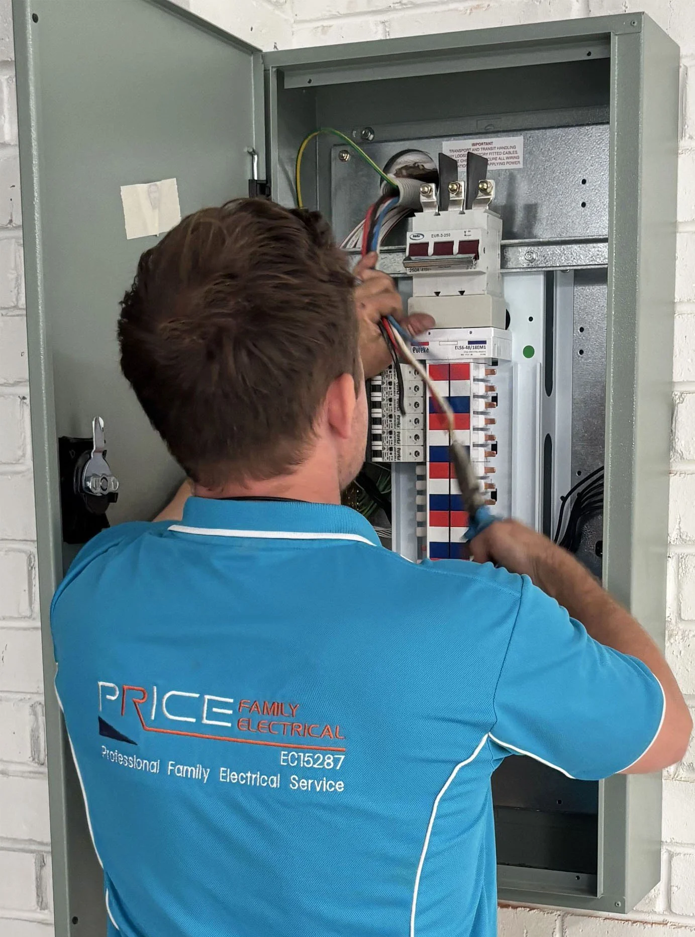 An electrician working on an open electrical panel, wearing a light blue shirt with the words 'Price Family Electrical' on the back.