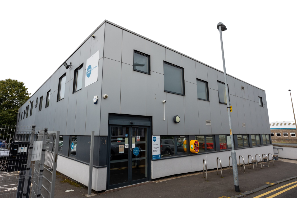 Grey commercial building with multiple windows and a sign that reads 'Gateshead'. Exterior includes a gated area, two automatic doors, and a parking lot with a yellow and red emergency call box.