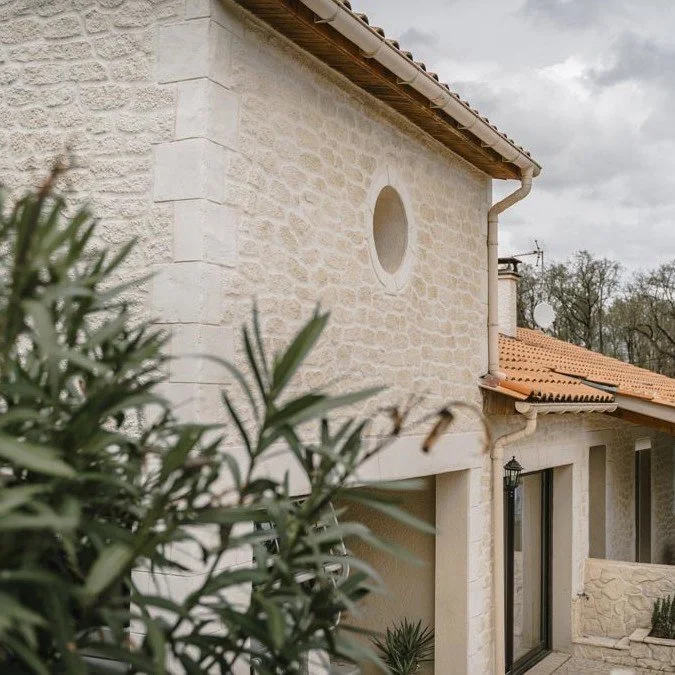 Façade d'une maison en pierre avec un mur en pierre beige, une fenêtre à cadre noir, un ancien style de lampadaire, et un toit en tuiles rouges, sous un ciel nuageux.