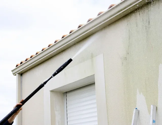 Une personne utilisant un pulvérisateur pour nettoyer ou faire de la peinture sur l'extérieur d'une maison, près d'une fenêtre.