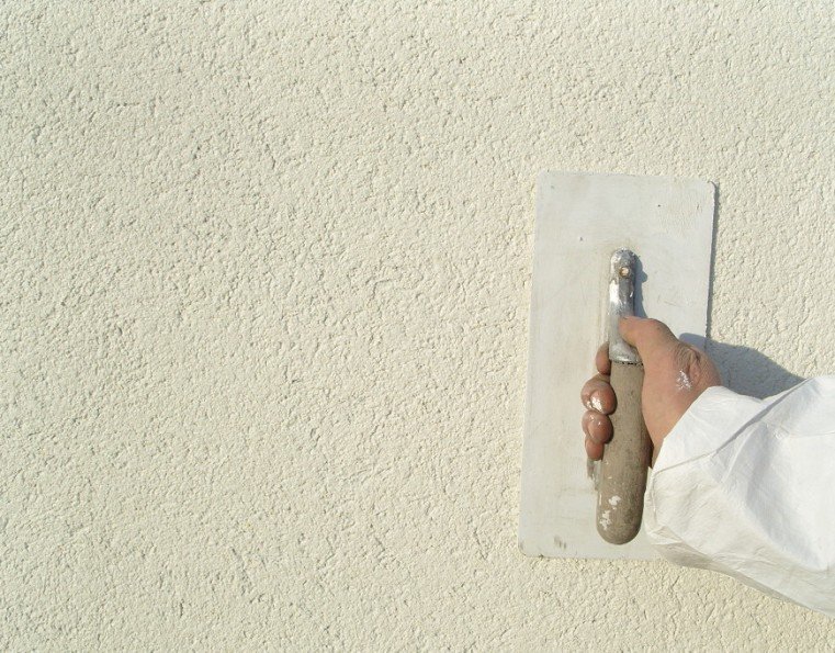 Une main tenant une truelle contre un mur en plâtre beige.