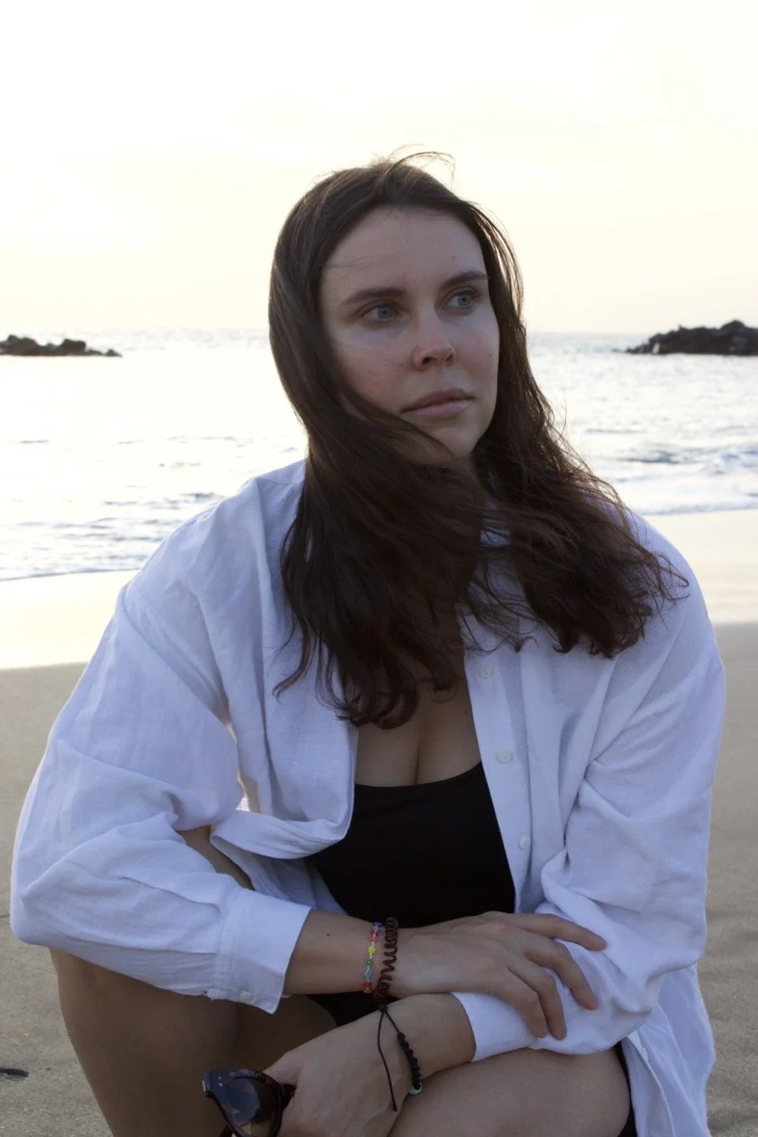 A close-up portrait of a woman on the beach at sunset, wearing a white shirt with hair blowing across her face.
