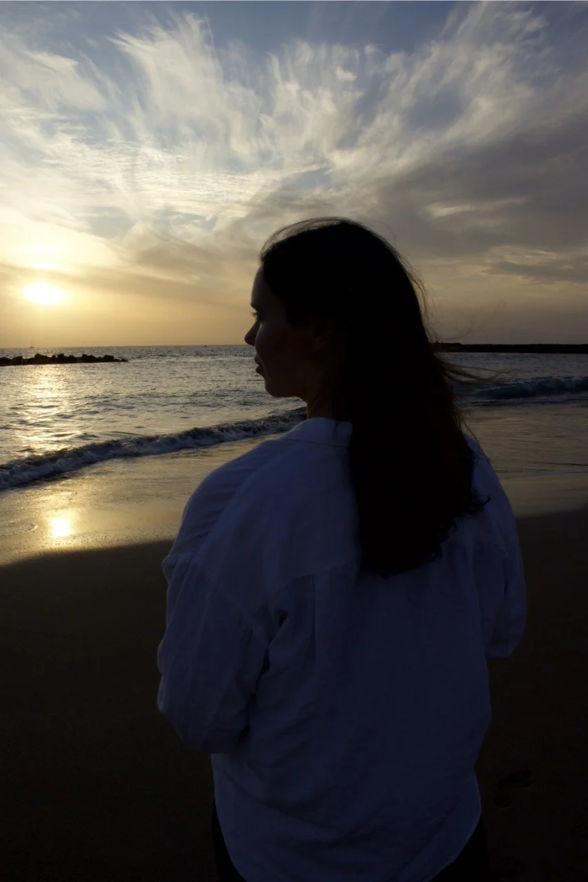 A silhouette of a woman in a white shirt facing the ocean as the sun sets behind scattered clouds.