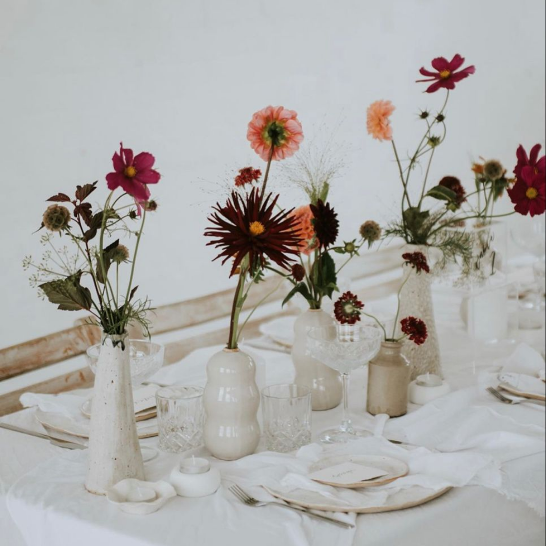 Mesa decorada con varios jarrones con flores variadas en tonos rosas y rojos, y cristalería fina, en un estilo elegante y minimalista.