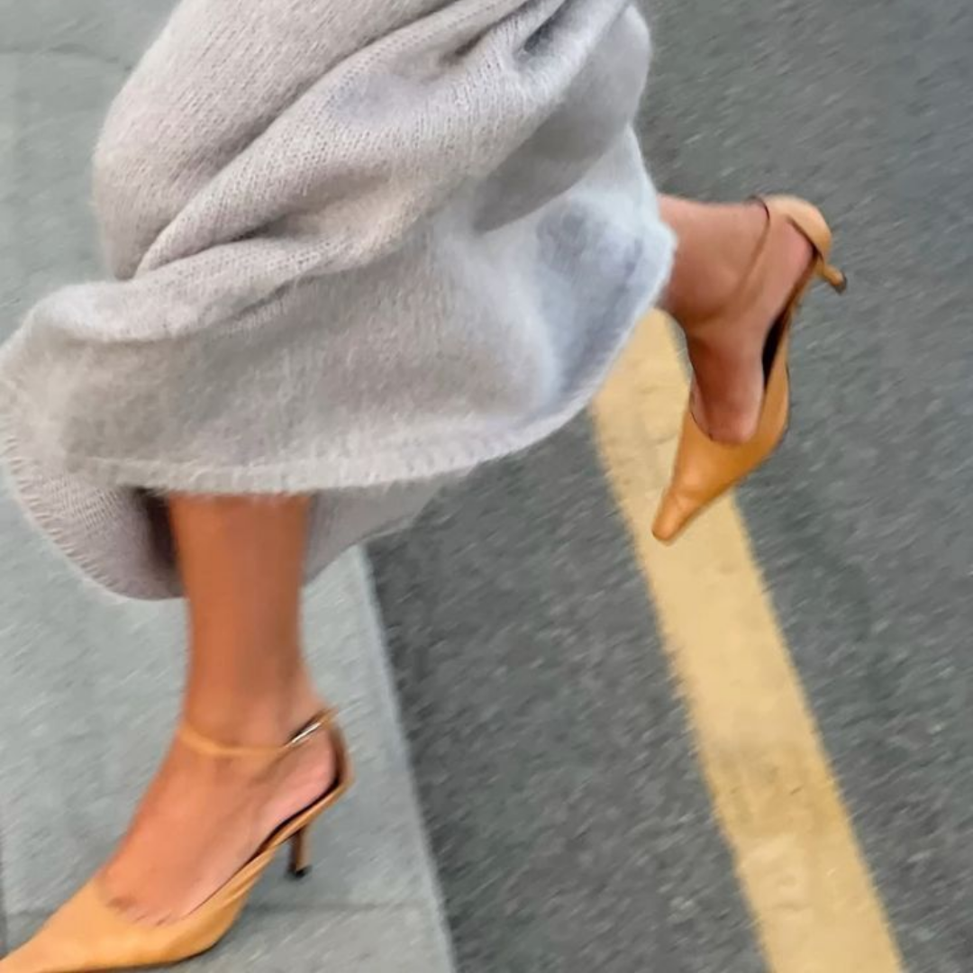 Mujer caminando con falda gris y tacones beige en la calle.