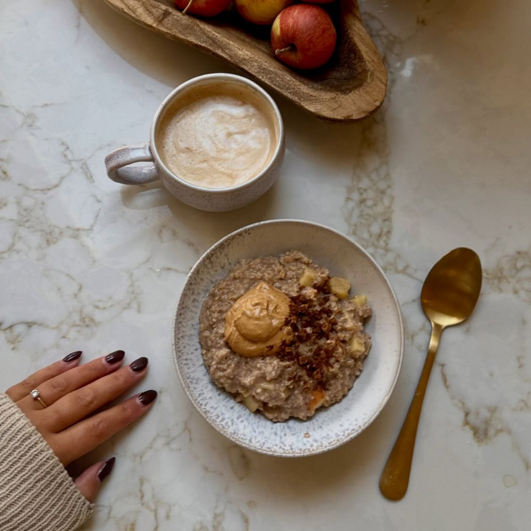 Desayuno con avena, manzana, café y una cucharada de mantequilla de maní en un plato, en una mesa de mármol, con mano de mujer y un plato de manzanas y un vaso de café.