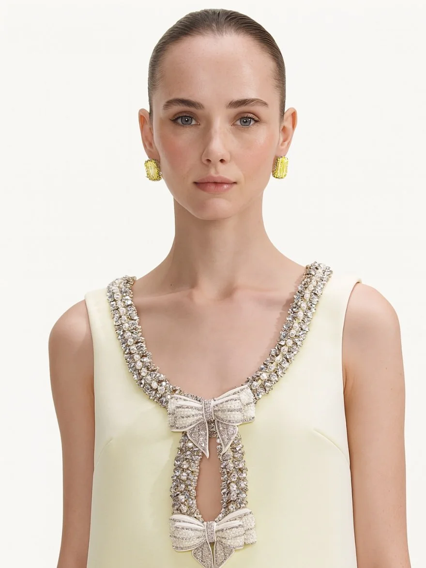 A woman with slicked-back hair wearing a sleeveless cream-colored dress decorated with rhinestones and bows at the neckline, and large yellow earrings.