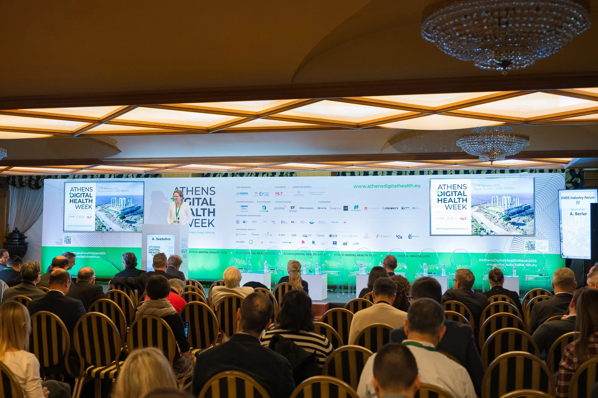 Audience attending the Athens Digital Health Week conference in a large conference room with a presenter on stage, multiple screens displaying event information, and chandeliers hanging from the ceiling.