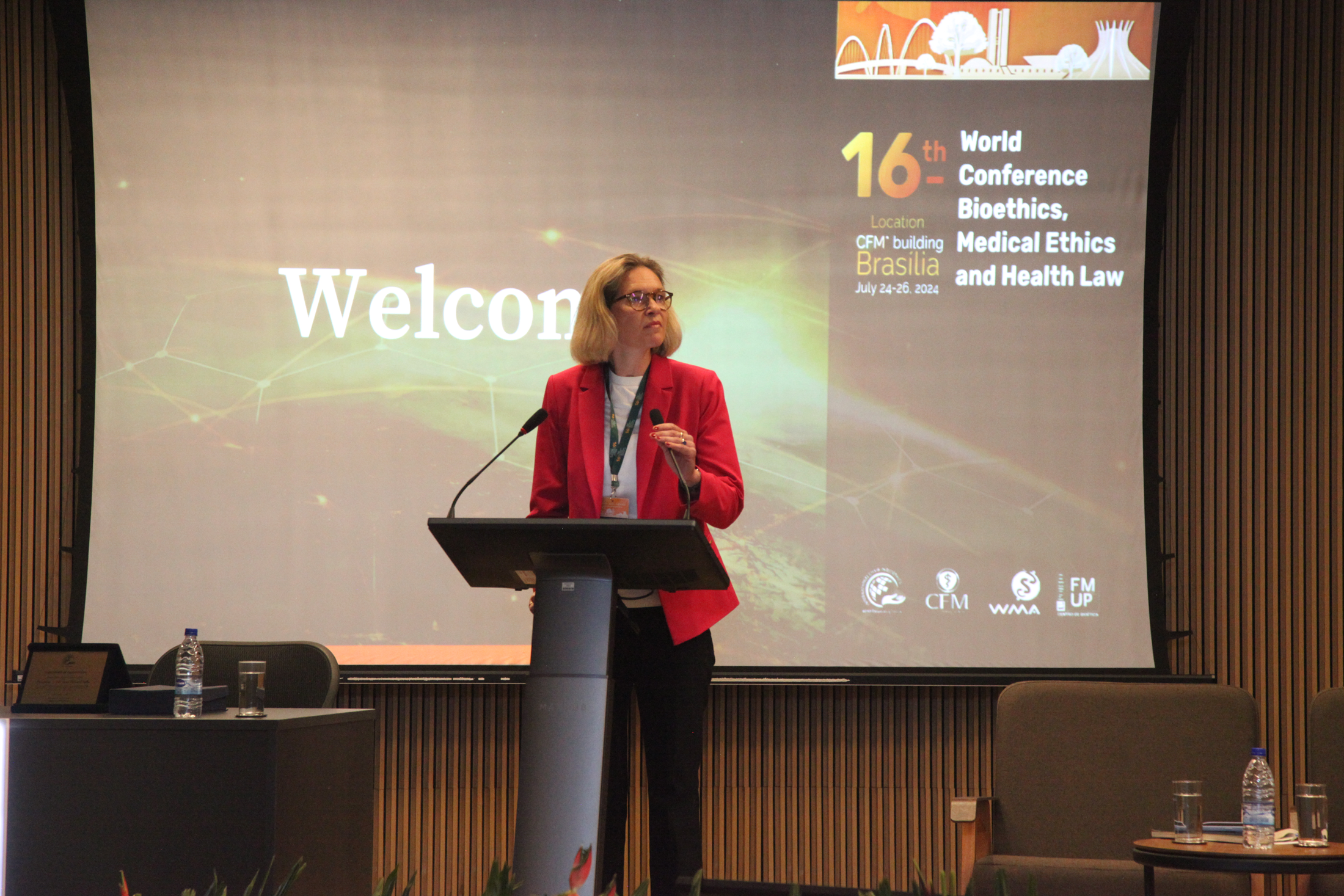 A woman with glasses and blond hair, wearing a red blazer, standing at a podium with a presentation screen behind her at a conference, with the screen displaying information about the 16th World Conference on Bioethics, Medical Ethics, and Health Law in Brasilia, July 24-26, 2024.